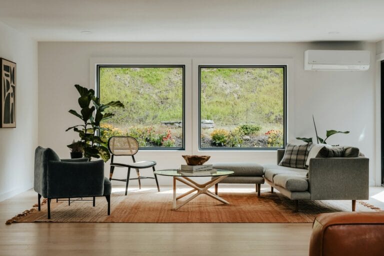 Bright living room with two large picture windows, a grey sofa, a wall-mounted split air conditioner and a green garden view outside