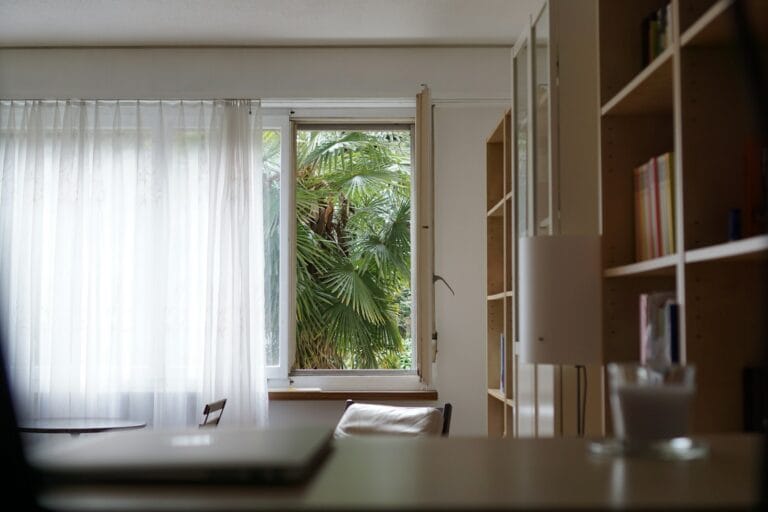 Room with sheer white curtains and an open window looking out onto palm leaves