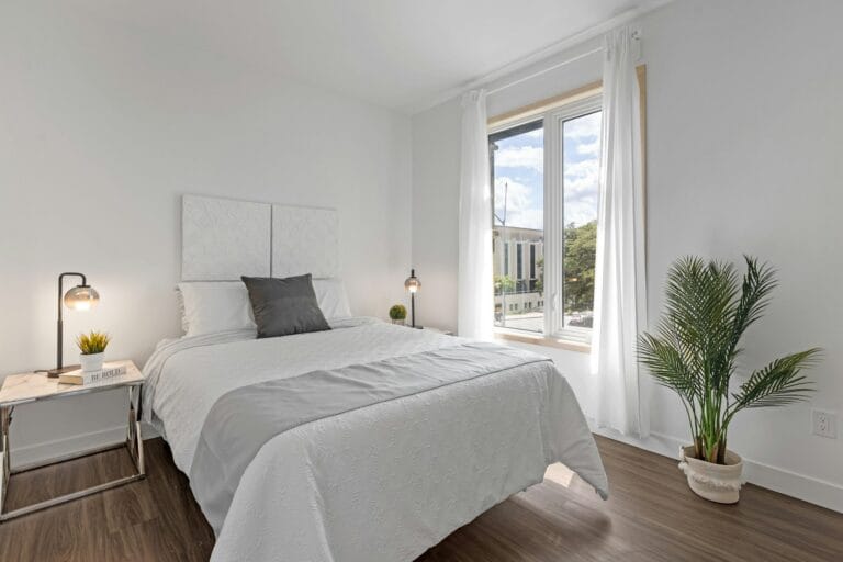 Bright bedroom with a double bed, a white-framed window and sheer white curtains, with a building and trees visible outside