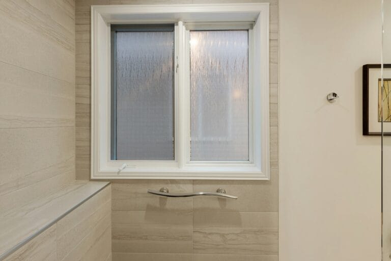 White-framed casement window with textured privacy glass above a chrome grab bar in a tiled bathroom