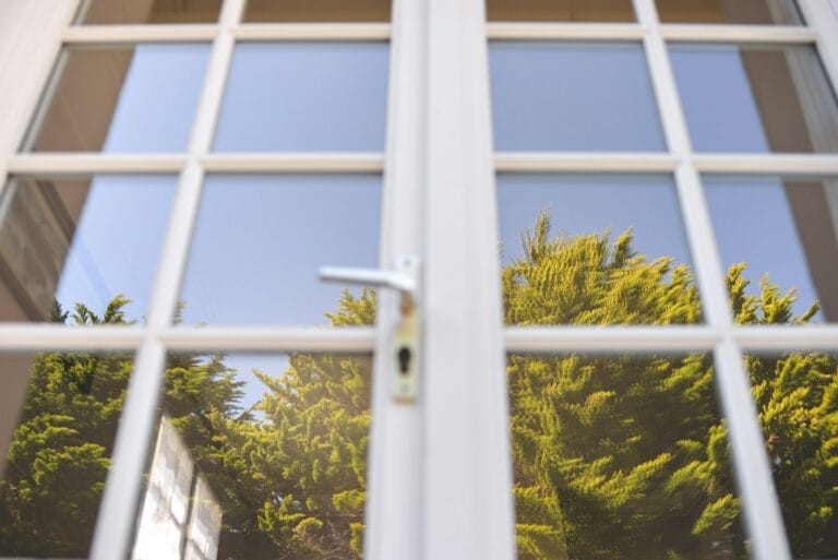 Close-up of white-framed glazed panes with grid bars and a lever handle, reflecting green conifer trees and a blue sky