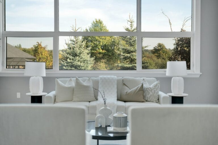 Bright living room with a white sofa and two table lamps in front of a wide white-framed window overlooking trees